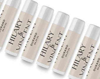 May include: A series of cylindrical lip balm tubes with white caps and beige labels. Each label features the black printed text HILARY & VINCENT, the name DURAND, and the date 04.27.24, arranged in a clean, minimalist sans-serif font style.