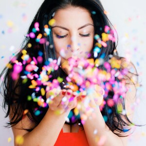 May include: A woman with long black hair blows confetti out of her hands. The confetti is multi-colored and creates a festive atmosphere.