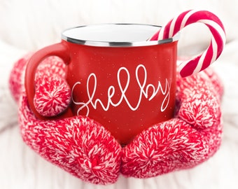 Peut inclure: Mug en émail rouge avec une inscription blanche qui dit "Shelby" et une canne en sucre qui dépasse du haut. Le mug est tenu par des mitaines en tricot rouge et blanc.