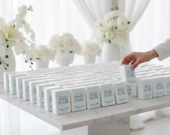 May include: A large collection of rectangular white tissue packets arranged in rows on a marble surface. Each packet features a light blue label with the text "FOR YOUR Happy TEARS" followed by "ANDREA + JAMES" and "05.11.24" printed in a dark blue script and serif font.