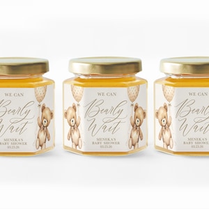 May include: Three small glass jars of honey with labels that say "We can Bearly Wait" with a teddy bear and balloons. The labels are decorated with a light brown and white polka dot pattern. The text on the labels is in a gold script font. The jars are on a white background.