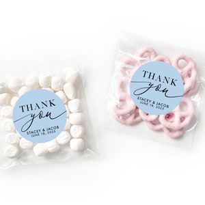 Peut inclure: Deux sachets en plastique transparent avec des autocollants ronds bleus et blancs qui disent "Merci" avec les noms Stacey & Jacob et la date du 18 juin 2025. Un sachet contient des guimauves blanches et l'autre contient des bretzels roses.