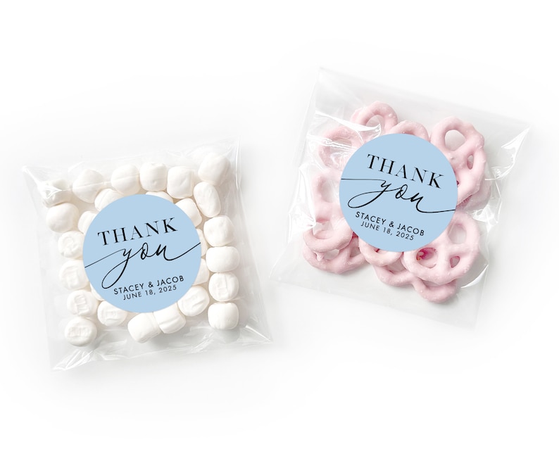 Peut inclure: Deux sachets en plastique transparent avec des autocollants ronds bleus et blancs qui disent "Merci" avec les noms Stacey & Jacob et la date du 18 juin 2025. Un sachet contient des guimauves blanches et l'autre contient des bretzels roses.