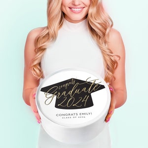 May include: A white round gift box with a black graduation cap design and gold lettering that says "Congrats Graduate 2024" and "Congrats Emily! Class of 2024".