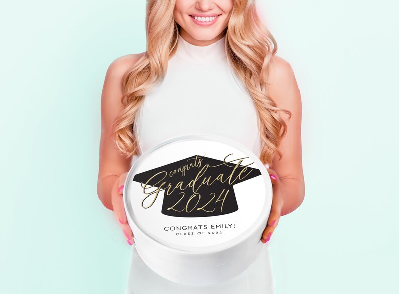 May include: A white round gift box with a black graduation cap design and gold lettering that says "Congrats Graduate 2024" and "Congrats Emily! Class of 2024".