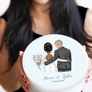 May include: A white round box with a blue background and a drawing of a bride and groom. The bride is wearing a white dress and the groom is wearing a black suit. The text "Imani & Tyler JOHNSON" is printed on the box.