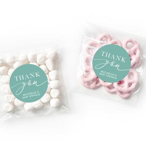 Peut inclure: Deux sacs en plastique transparent avec des autocollants de remerciement. L'autocollant est un cercle turquoise avec du texte blanc qui dit "Merci" avec une police d'&eacute;criture cursive "vous". Le texte en dessous dit "Baby Sprinkle de Michelle". Un sac contient des guimauves blanches et l'autre contient des bouch&eacute;es de bretzels roses.