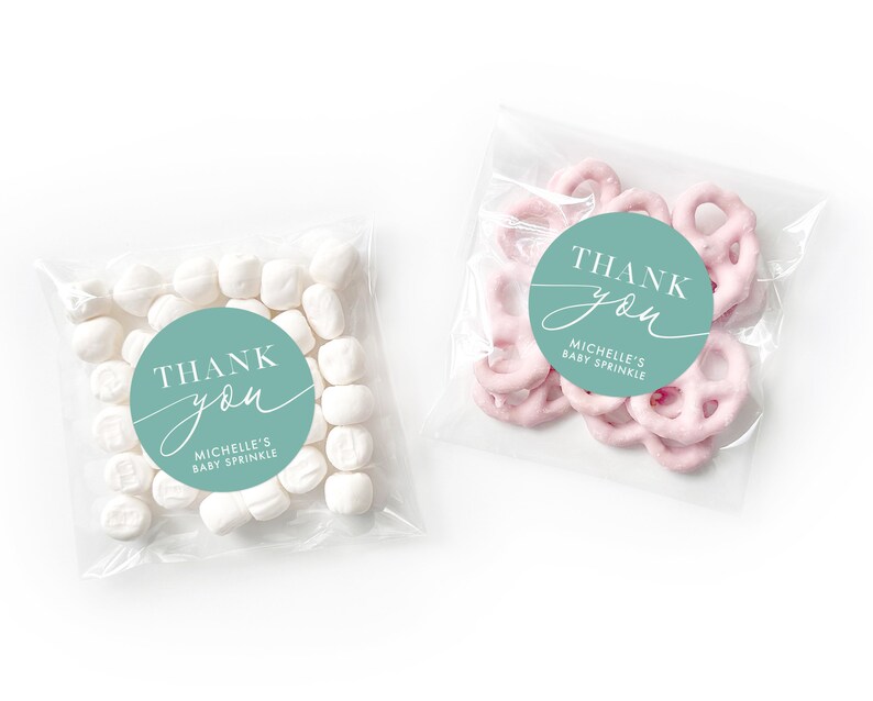 Peut inclure: Deux sacs en plastique transparent avec des autocollants de remerciement. L'autocollant est un cercle turquoise avec du texte blanc qui dit "Merci" avec une police d'&eacute;criture cursive "vous". Le texte en dessous dit "Baby Sprinkle de Michelle". Un sac contient des guimauves blanches et l'autre contient des bouch&eacute;es de bretzels roses.