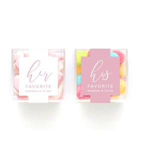 May include: Two clear plastic containers filled with candy. One container has pink and white marshmallows and a white label with the text "her favorite Hannah & Tyler". The other container has colorful candies and a pink label with the text "his favorite Hannah & Tyler".