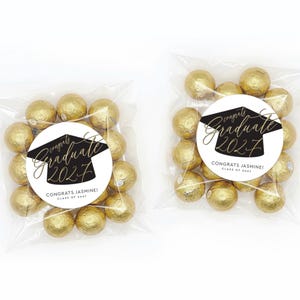 May include: Two clear cellophane bags filled with gold-wrapped candies. Each bag has a round sticker with the text "Congrats Jasmine! Graduate 2027" in black and gold script and a graduation cap illustration.