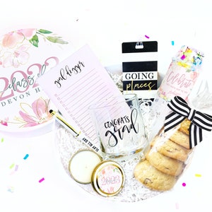 May include: A graduation gift box filled with a variety of items, including a pink and white floral round box with the text "Class of 2022 Devon H", a pink notepad with the text "goal digger", a clear glass mug with the text "Congrats Grad", a black and white striped gift tag with the text "Going Places", a clear plastic bag with confetti and the text "Celebrate Say hats off to you Graduate", a candle in a gold tin with a floral design and the text "2023", and a stack of chocolate chip cookies wrapped in clear plastic with a black and white striped ribbon.