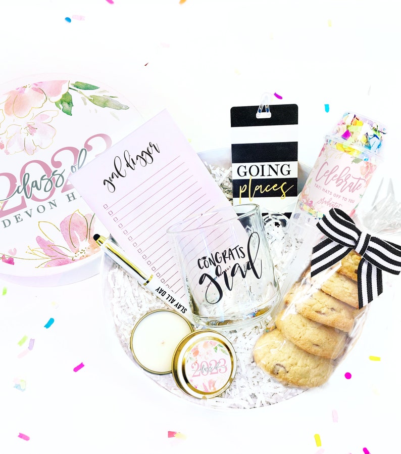 May include: A graduation gift box filled with a variety of items, including a pink and white floral round box with the text "Class of 2022 Devon H", a pink notepad with the text "goal digger", a clear glass mug with the text "Congrats Grad", a black and white striped gift tag with the text "Going Places", a clear plastic bag with confetti and the text "Celebrate Say hats off to you Graduate", a candle in a gold tin with a floral design and the text "2023", and a stack of chocolate chip cookies wrapped in clear plastic with a black and white striped ribbon.