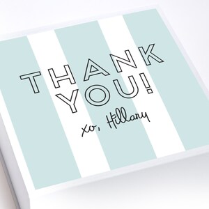 May include: A white gift box with blue and white stripes. The box has the text "THANK YOU!" in black outlined letters. The text "xo, Hillary" is written in black cursive below the main text.