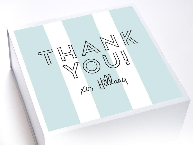 May include: A white gift box with blue and white stripes. The box has the text "THANK YOU!" in black outlined letters. The text "xo, Hillary" is written in black cursive below the main text.