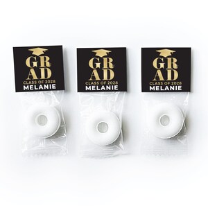 May include: Three clear plastic bags containing white ring-shaped candies. Each bag has a black header card with a gold graduation cap icon, the text GRAD, CLASS OF 2028, and the name MELANIE printed in white and gold lettering.