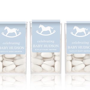 May include: Three clear containers of white mints with blue and white labels. The labels feature a white rocking horse and the text "celebrating Baby Hudson Kayla's Baby Shower".