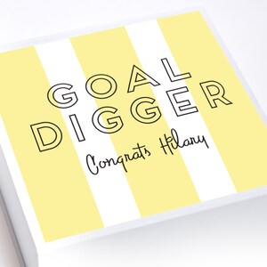 May include: A white gift box with yellow and white stripes. The box has the text "GOAL DIGGER" in black outlined letters and the text "Congrats Hilary" in black cursive writing.