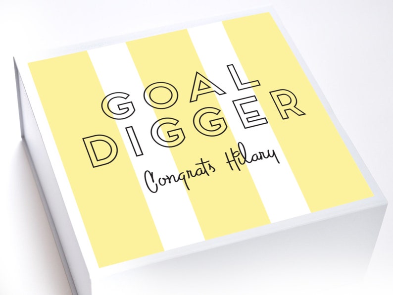 May include: A white gift box with yellow and white stripes. The box has the text "GOAL DIGGER" in black outlined letters and the text "Congrats Hilary" in black cursive writing.