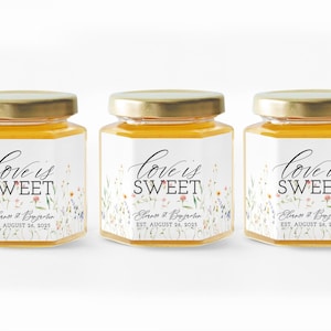 May include: Three hexagonal glass jars filled with golden honey, each topped with a gold metal lid. The labels feature a watercolor wildflower design, the text "love is SWEET," the names "Eleanor & Benjamin," and "EST. AUGUST 26, 2025."