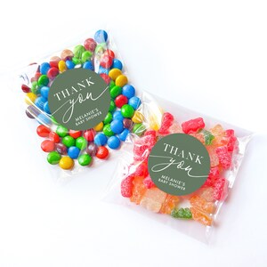 Peut inclure: Deux sacs en plastique transparents remplis de bonbons color&eacute;s. Un sac contient des bonbons g&eacute;lifi&eacute;s verts et rouges, l'autre des M&Ms multicolores. Les deux sacs ont des autocollants ronds verts avec du texte blanc qui dit "Merci" et "Baby Shower de M&eacute;lanie".