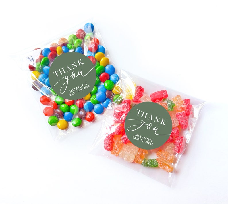 Peut inclure: Deux sacs en plastique transparents remplis de bonbons color&eacute;s. Un sac contient des bonbons g&eacute;lifi&eacute;s verts et rouges, l'autre des M&Ms multicolores. Les deux sacs ont des autocollants ronds verts avec du texte blanc qui dit "Merci" et "Baby Shower de M&eacute;lanie".