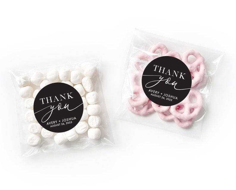 Peut inclure: Deux sacs en plastique transparents remplis de cadeaux de mariage. Les sacs ont des autocollants ronds noirs et blancs qui disent "Merci" avec les noms du couple et la date du mariage. Un sac contient des guimauves blanches et l'autre contient des bretzels roses.