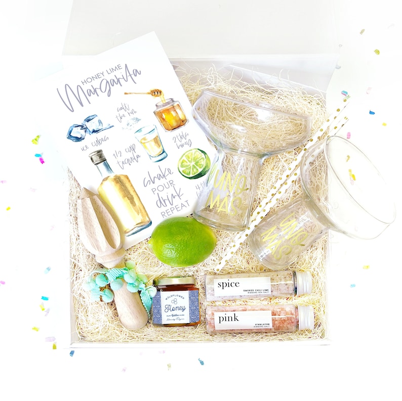 May include: A gift box filled with items for making margaritas. The box contains a recipe card, a bottle of tequila, a lime, a honey jar, a lime squeezer, a margarita glass, two shot glasses, two straws, and two small jars of salt. The shot glasses and straws are decorated with the word "Margarita".