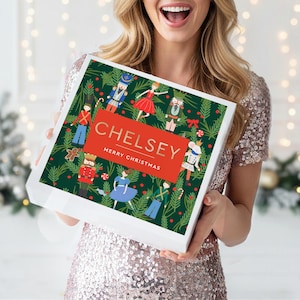 May include: A square white gift box featuring a dark green background with a pattern of nutcrackers, ballerinas, and pine branches. A central red rectangle displays the name CHELSEY in gold letters above the text MERRY CHRISTMAS.