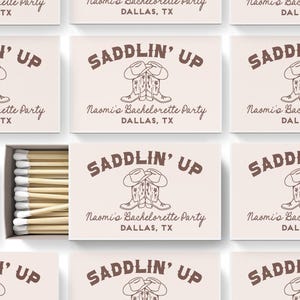 May include: A set of matchboxes with a brown and white design. The matchboxes have a graphic of two cowboy boots with hats on top. The text "SADDLIN' UP" is printed in brown on the matchboxes. The text "Naomi's Bachelorette Party" and "Dallas, TX" is also printed on the matchboxes.