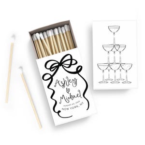 May include: White matchbox with black calligraphy that reads "Ashley & Michael August 05, 2023 New York, NY" and a black ribbon illustration. The matchbox is open and shows wooden matches with white tips.