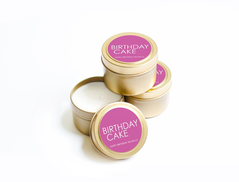 以下が含まれることがあります： ピンクのラベルが付いた金色のキャンドル缶が4つ。ラベルには「Birthday Cake」と「Happy Birthday Michelle」と書かれています。