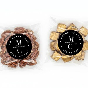 May include: Two clear cellophane bags, each containing treats. One bag holds a cluster of pecans, the other contains gold-wrapped chocolates. Both bags feature a black circular sticker with white text that reads "CONGRATS GRAD" and "CLASS OF 2027".