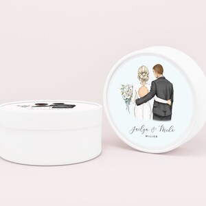 May include: White round box with a blue background and a watercolor illustration of a bride and groom. The text "Jaelya & Mike MILLER" is printed below the illustration.