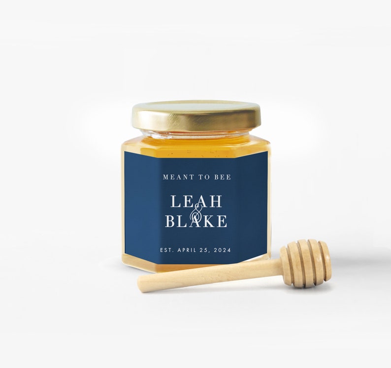 Pu&ograve; includere: Un piccolo barattolo di miele in vetro con coperchio dorato ed etichetta blu navy. L'etichetta recita "MEANT TO BEE" e "LEAH & BLAKE" con la data "EST. APRIL 25, 2024". Un cucchiaino di miele in legno &egrave; appoggiato accanto al barattolo.