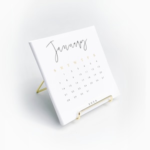 May include: A gold metal desk calendar stand with a white calendar insert for the month of January 2024.