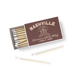 May include: A brown matchbox with a white label that says "NASHVILLE" in a white outline. The label also has a drawing of two cowboy boots with spurs and a floral design. The text "Adriana's 30th Birthday" and "JUNE 5, 2026" is also on the label. The matchbox is open and there are several matches inside.