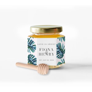 Pu&ograve; includere: Un piccolo barattolo di miele in vetro con coperchio dorato e un cucchiaio di miele in legno. Il barattolo ha un'etichetta con il testo "Love is Sweet" e i nomi "Fiona & Henry" con la data "Est. July 25, 2020". L'etichetta ha uno sfondo rosa con foglie tropicali verdi e blu.