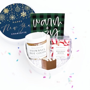May include: A white gift box filled with hot cocoa mix, a chocolate spoon, and holiday mints. The box includes a navy blue lid with gold snowflakes and the text "Happy New Year Charlotte." A green and black plaid card reads "Warm + Cozy."