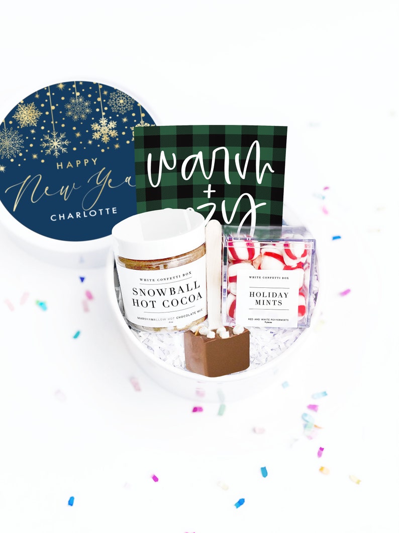 May include: A white gift box filled with hot cocoa mix, a chocolate spoon, and holiday mints. The box includes a navy blue lid with gold snowflakes and the text "Happy New Year Charlotte." A green and black plaid card reads "Warm + Cozy."