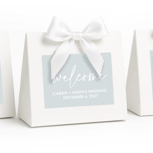 May include: A white triangular gift box featuring a light blue rectangular label with the white text welcome, CARRIE + AIDEN'S WEDDING, and DECEMBER 6, 2027. The box is topped with a white satin ribbon tied into a decorative bow at the center.