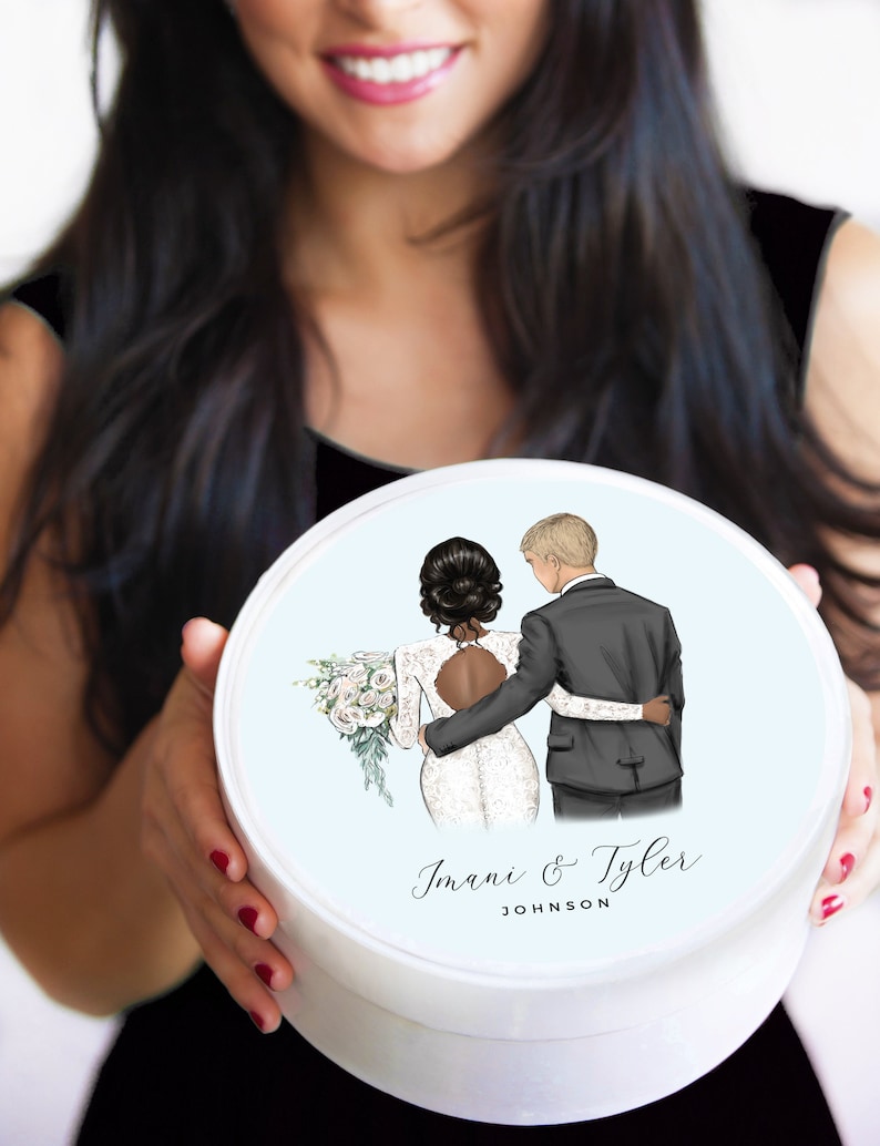 May include: A white round box with a blue background and a cartoon illustration of a bride and groom. The bride is wearing a white dress and the groom is wearing a black suit. The text "Imani & Tyler JOHNSON" is printed on the box.