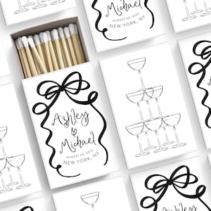 May include: White matchboxes with black and white illustrations of champagne glasses and a bow. The text on the matchboxes reads "Ashley & Michael" and includes the date and location of the wedding.