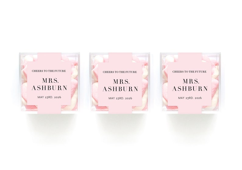 May include: Three clear boxes filled with pink and white marshmallows. Each box has a pink label with the text "CHEERS TO THE FUTURE MRS. ASHBURN MAY 23RD, 2026".