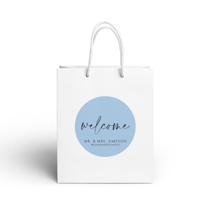 A white paper shopping bag with a blue circular logo and the text "welcome Mr. & Mrs. Simpson" printed on it.