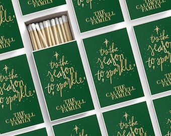 Puede incluir: Cajas de cerillas verdes con letras doradas que dicen "tis the season to sparkle" y "The Caldwell Family". Una caja está abierta, revelando cerillas de madera con puntas blancas. Las cajas están dispuestas sobre una superficie blanca.