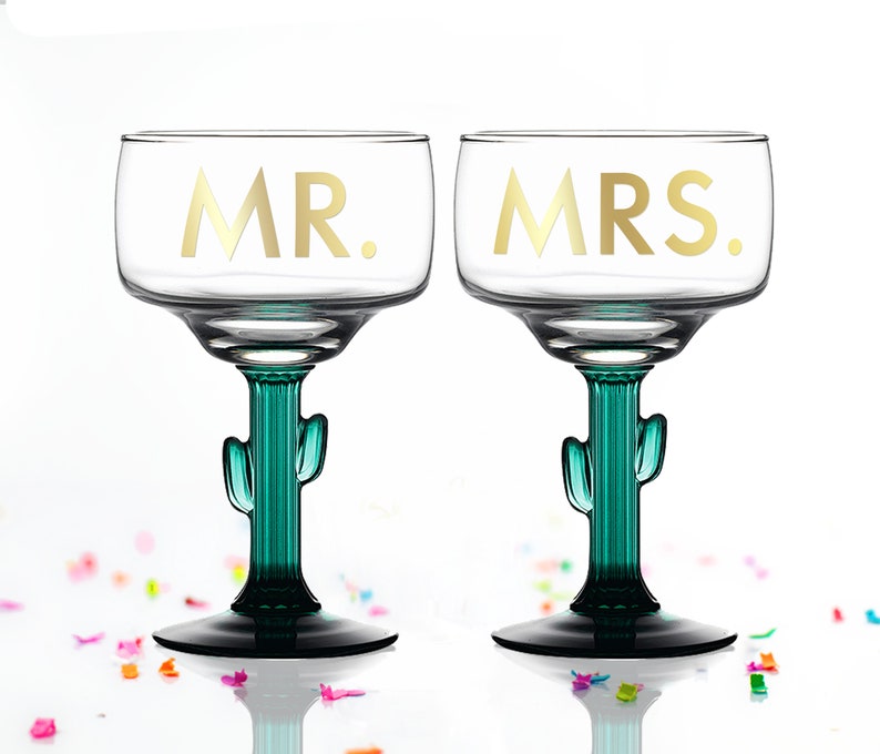 May include: Two clear glass margarita glasses with gold lettering that says "MR." and "MRS." The glasses have green cactus-shaped stems and bases.