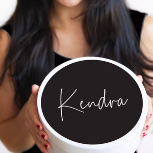 K&ouml;nnte beinhalten: Eine wei&szlig;e runde Geschenkbox mit einem schwarzen Deckel. Der Deckel tr&auml;gt den Namen "Kendra" in wei&szlig;er Kursivschrift.