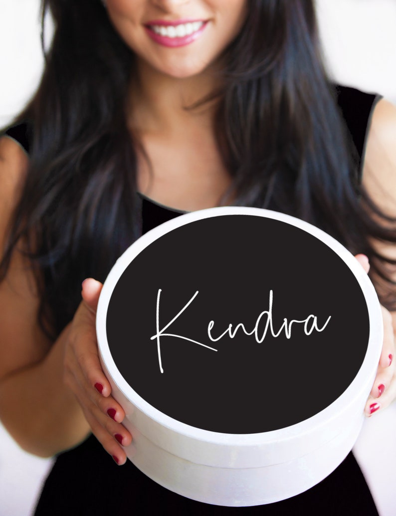 K&ouml;nnte beinhalten: Eine wei&szlig;e runde Geschenkbox mit einem schwarzen Deckel. Der Deckel tr&auml;gt den Namen "Kendra" in wei&szlig;er Kursivschrift.