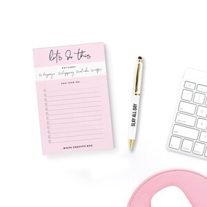 May include: Pink notepad with the text "let's do this" and a checklist. The checklist includes the text "but first:  " and has checkboxes for "yoga", "blogging", "outtake", and "coffee". The notepad also includes the text "and then do:" and has a list of blank checkboxes. The notepad is on a white surface with a white pen and a white keyboard.