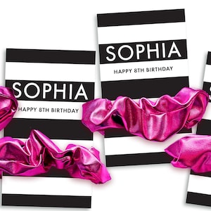 May include: Cards with black and white stripes, the name "SOPHIA" in large white letters, and the text "HAPPY 8TH BIRTHDAY". Bright pink scrunchies are in the foreground. The cards and scrunchies are arranged on a white surface.